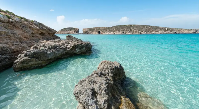 Crystal-clear shallow water over rocks at Comino's Blue Lagoon, with the limestone coastline behind